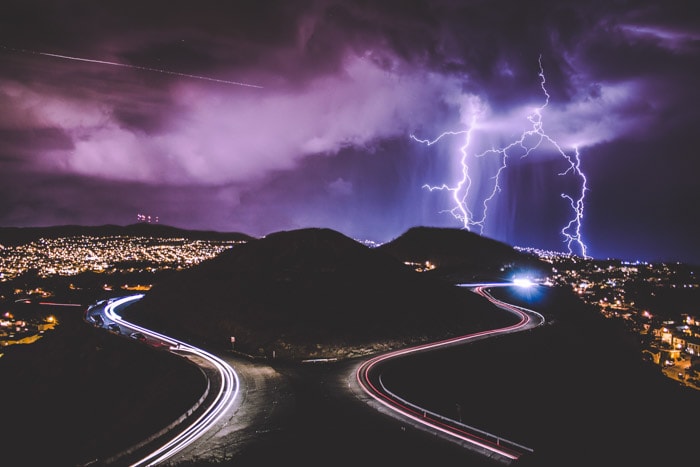 Magnificent overhead shot of a two roads with colored light trails bypassing a hill, fantastic purple skies and lightning striking above